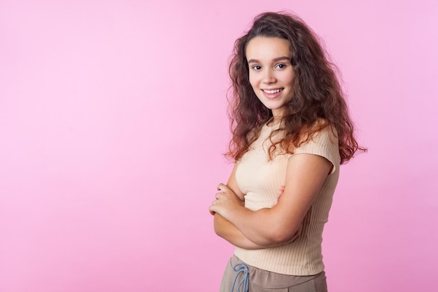 Porträt eines fröhlichen, selbstbewussten Teenager-Mädchens mit langen, lockigen, brünetten Haaren in beigefarbener Kleidung im lässigen Stil, das mit gekreuzten Händen steht und ein attraktives Lächeln im Studio aufnimmt, isoliert auf rosa Hintergrund