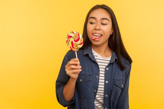 Porträt eines fröhlichen, sorglosen Mädchens im Jeanshemd, das mit herausgestreckter Zunge steht, träumt und versucht ist, Lollipop zu probieren, runde, süße, zuckerhaltige Regenbogenbonbons. Studioaufnahme im Innenbereich isoliert auf gelbem Hintergrund