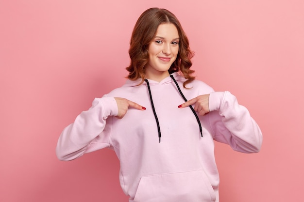 Porträt eines fröhlichen Teenager-Mädchens mit lockigen Haaren, das lächelt und mit dem Finger auf sich selbst zeigt, wenn es mit Stolz und Freude in die Kamera schaut Innenstudioaufnahme isoliert auf rosa Hintergrund