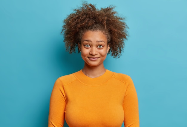 Porträt eines fröhlichen, überraschten tausendjährigen Mädchens mit lockigem, buschigem Haar hat sich gefragt, ob der Ausdruck einen orangefarbenen Pullover trägt