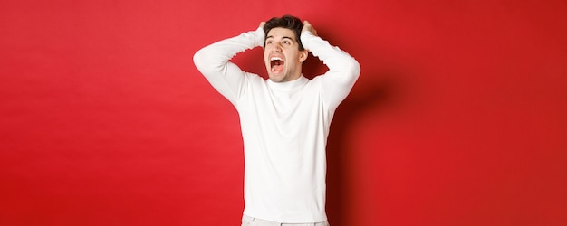 Porträt eines frustrierten Mannes im weißen Pullover, der in Panik schreit und auf die obere linke Ecke schaut, hat ein schreckliches Problem und steht auf rotem Hintergrund