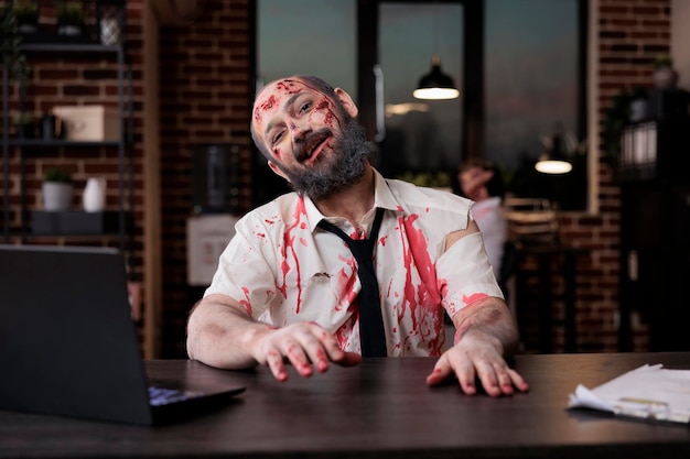 Foto porträt eines gefährlichen zombie-mannes, der am schreibtisch sitzt und im firmenbüro gruselig und erschreckend aussieht. gehirnfressendes grausames monster mit blutigen wunden, das am laptop arbeitet, schrecklicher besessener teufel.