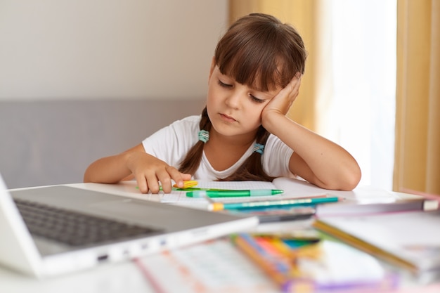 Porträt eines gelangweilten Schulmädchens, das weißes T-Shirt trägt, am Tisch sitzt, vor dem Notebook, traurigen Gesichtsausdruck hat, müde ist, Hausaufgaben zu machen, Fernunterricht.