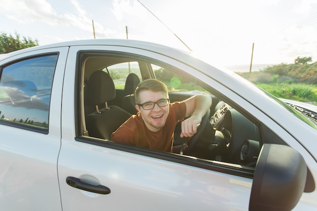 Porträt eines glücklich lächelnden jungen Mannes, Käufer, der in seinem neuen Auto sitzt und Schlüssel außerhalb des Händlerbüros zeigt.