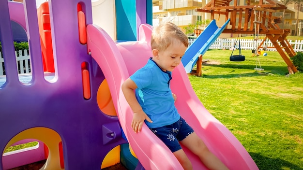 Porträt eines glücklich lächelnden Kleinkindjungen, der auf einer bunten Plastikrutsche auf dem Kinderspielplatz im Park reitet