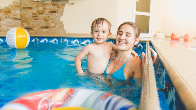 Porträt eines glücklich lächelnden Kleinkindjungen, der mit aufblasbarem buntem Wasserball mit Mutter im Innenpool spielt