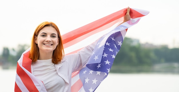 Porträt eines glücklich lächelnden rothaarigen Mädchens mit USA-Nationalflagge auf ihren Schultern. Positive junge Frau, die den Unabhängigkeitstag der Vereinigten Staaten feiert.