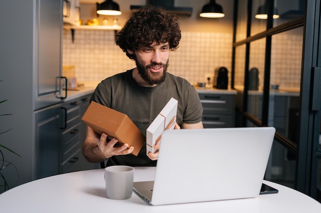 Porträt eines glücklich überraschten jungen lockigen Mannes, der während eines Videoanrufs auf einem Laptop am Tisch in der Küche eine Geschenkbox mit Geschenk öffnet