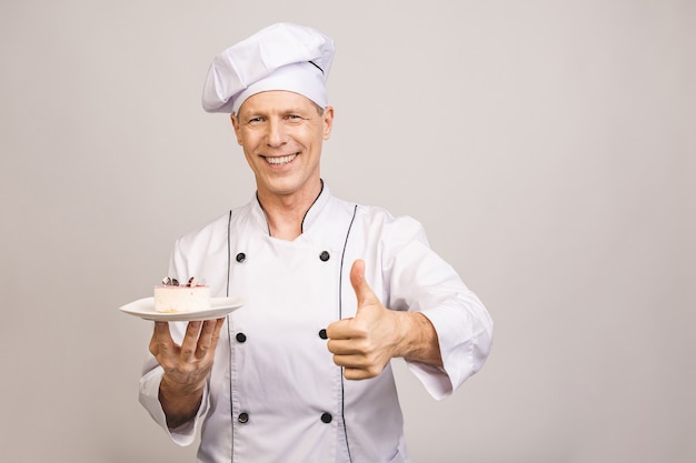 Porträt eines glücklichen älteren männlichen Kochs gekleidet in der Uniform, die Platte mit Stück Kuchen Daumen hoch hält.