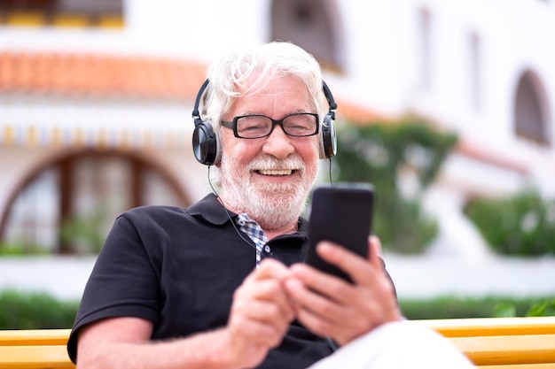 Porträt eines glücklichen älteren weißhaarigen Mannes mit Bart, der auf einer Bank sitzt und Musik hört