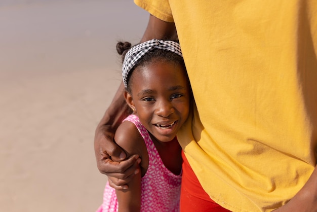 Porträt eines glücklichen afroamerikanischen Mädchens, das seinen jungen Vater umarmt, während es an einem sonnigen Tag am Strand steht