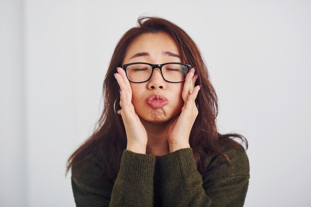 Porträt eines glücklichen asiatischen jungen Mädchens in Brillen, das drinnen im Studio vor weißem Hintergrund steht.