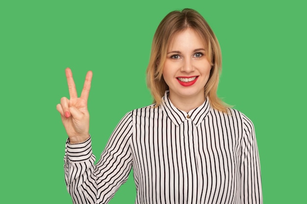 Porträt eines glücklichen, attraktiven Mädchens in gestreifter Bluse, das mit zwei Fingern den Sieg oder die Friedensgeste zeigt und mit einem charmanten, zahnigen Lächeln in die Kamera blickt, Studioaufnahme isoliert auf grünem Hintergrund