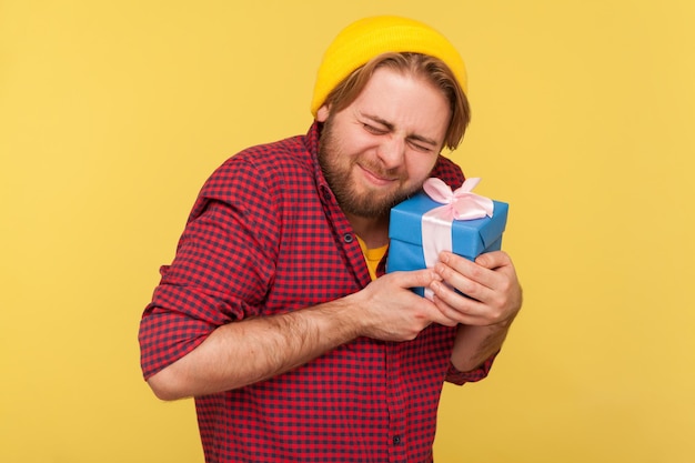 Porträt eines glücklichen bärtigen Hipster-Typen mit Beanie-Hut und kariertem Hemd, der steht und die Geschenkbox hält und sie umarmt lang erwartetes Geschenk Indoor-Studioaufnahme isoliert auf gelbem Hintergrund