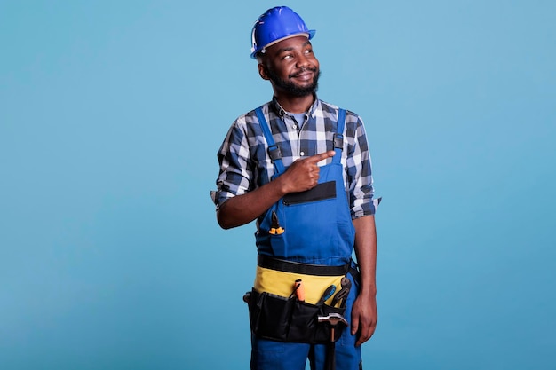 Porträt eines glücklichen Bauarbeiters bei der Arbeit mit Schutzhelm, während er mit dem Finger zur Seite zeigt. Tragender Gürtel des Elektrikers mit Werkzeugen in der Atelieraufnahme gegen blauen Hintergrund.