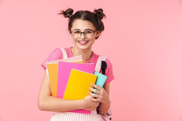 Porträt eines glücklichen, charmanten Mädchens, das mit Schulheften posiert und über rosafarbener Wand isoliert lächelt