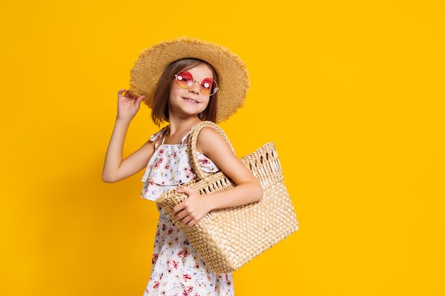 Porträt eines glücklichen, fröhlichen Mädchens in Sommerhut-Sonnenbrille-Strohbeutel über gelbem Hintergrund