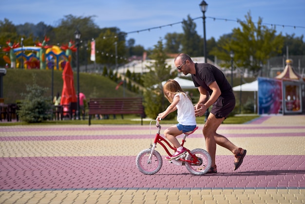 Porträt eines glücklichen fürsorglichen Vaters, der seine kleine hübsche Tochter unterrichtet, die Fahrrad in einem grünen Park fährt und in voller Länge lächelt