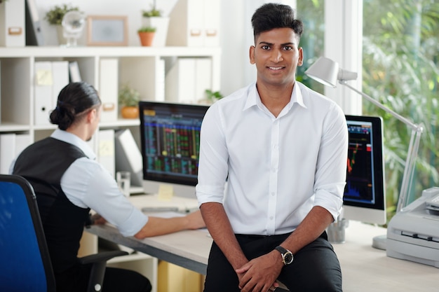 Porträt eines glücklichen, gutaussehenden jungen indischen Börsenmaklers, der sich auf den Bürotisch stützt und in die Kamera lächelt