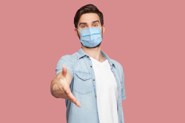 Porträt eines glücklichen, gutaussehenden jungen Mannes mit chirurgischer medizinischer Maske in blauem Hemd, der die Kamera mit einem Lächeln betrachtet und der Begrüßung die Hand gibt. Indoor-Studioaufnahme, isoliert auf rosa Hintergrund.