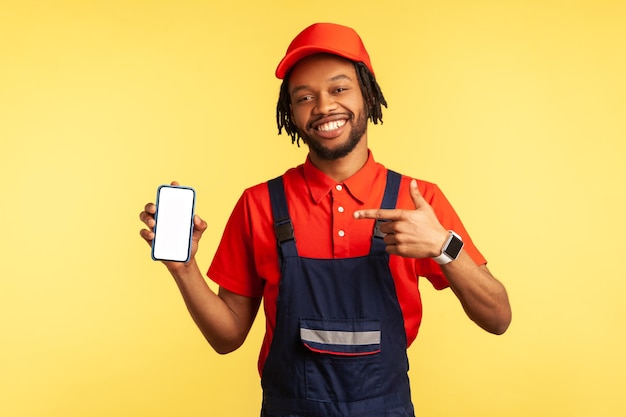 Porträt eines glücklichen Handwerkers in Uniform, der auf ein Smartphone mit leerem Bildschirm zeigt, der für neue Anwendungen in der Dienstleistungsbranche wirbt Studioaufnahme isoliert auf gelbem Hintergrund