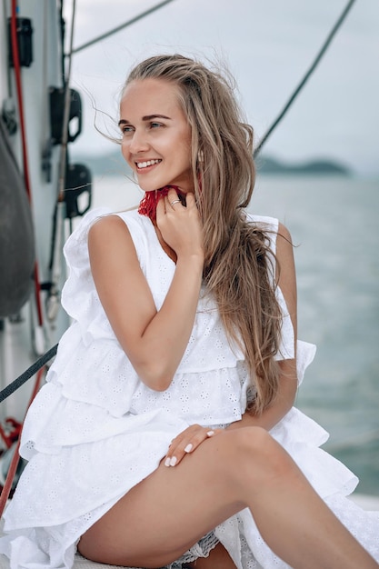 Porträt eines glücklichen hübschen Mädchens mit weißem Kleid, langen roten Ohrringen und langen lockigen blonden Haaren, das im Sommer auf der Yacht sitzt. Blick in die Kamera mit einem zahnigen Lächeln.