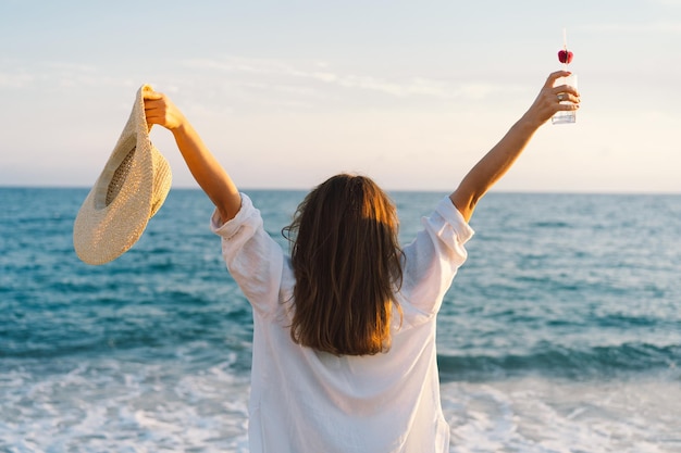 Porträt eines glücklichen jungen Mädchens in einem Strohhut mit einem Cocktail in der Hand auf einem Meereshintergrund