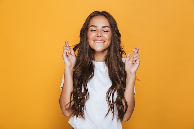 Porträt eines glücklichen jungen Mädchens mit langen brünetten Haaren, die über gelber Wand stehen und Daumen drücken für Glück