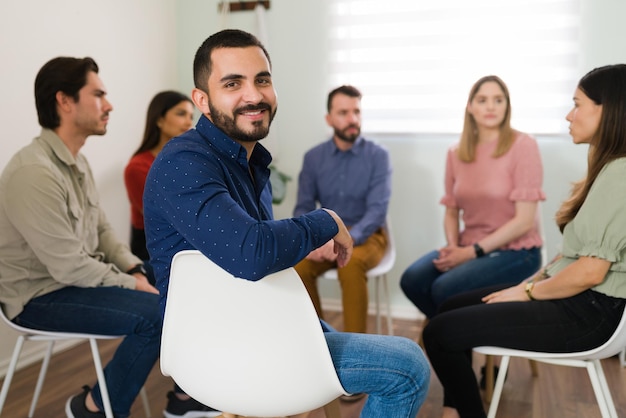 Porträt eines glücklichen jungen Mannes, der lächelt und Blickkontakt herstellt, während er während eines Treffens einer Selbsthilfegruppe sitzt. Neues Mitglied einer Aggressionsgruppe