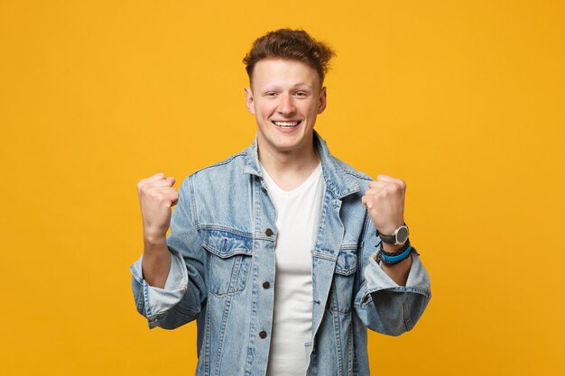 Porträt eines glücklichen jungen Mannes in Denim-Freizeitkleidung, der die Fäuste ballt, wie der Gewinner, isoliert auf gelb-orangefarbenem Wandhintergrund im Studio. Menschen aufrichtige Emotionen, Lifestyle-Konzept. Mock-up-Kopienbereich.