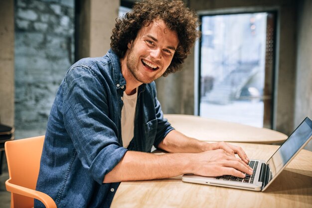 Porträt eines glücklichen jungen Mannes mit lockigem Haar, der Laptop zum Arbeiten und Surfen online verwendet Intelligenter kaukasischer Mann, der Nachrichten liest, die auf seinem Laptop-Computer im modernen Büro tippen Unternehmen und Menschen