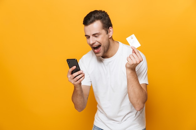 Porträt eines glücklichen jungen Mannes mit T-Shirt, der isoliert über gelber Wand steht, mit Handy, Plastikkreditkarte zeigt