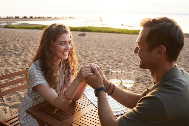 Porträt eines glücklichen jungen Paares, das sich beim Picknick im Freien an den Händen hält, während es ein romantisches Date am Strand genießt...