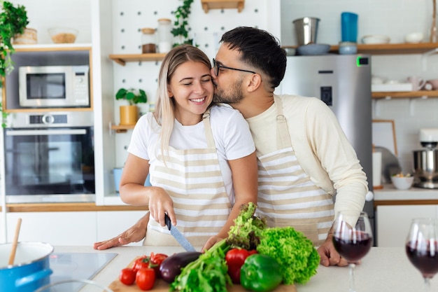 Porträt eines glücklichen jungen Paares, das zu Hause zusammen in der Küche kocht. Attraktive junge Frau und ein gutaussehender Mann genießen es, Zeit miteinander zu verbringen, während sie auf einer hellen, modernen Küche stehen