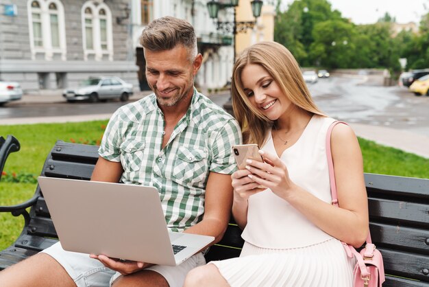 Porträt eines glücklichen jungen Paares in Sommerkleidung, das Kreditkarte hält und Laptop benutzt, während es auf der Bank in der Stadtstraße sitzt