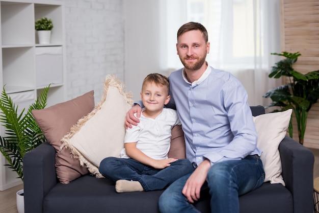Porträt eines glücklichen jungen Vaters mit kleinem Sohn, der zu Hause auf dem Sofa sitzt