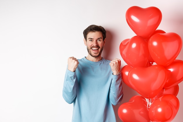 Porträt eines glücklichen Kerls, der den Tag der Liebenden feiert, in der Nähe des roten Herzballons des Valentinsgrußes steht und jubelt, eine Ja-Geste macht und in die Kamera lächelt, auf weißem Hintergrund steht.