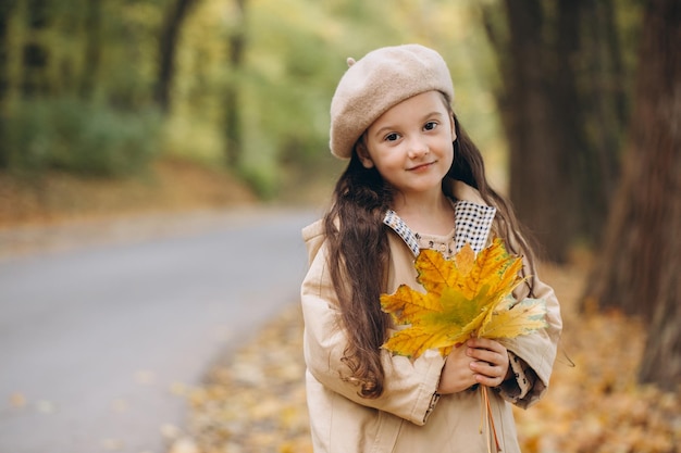 Porträt eines glücklichen kleinen Mädchens in beigem Mantel und Baskenmütze, das gelbe Ahornblätter hält und Zeit im Herbstpark verbringt