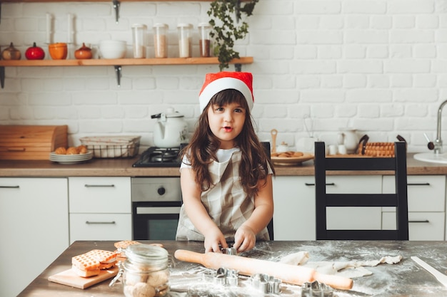 Porträt eines glücklichen kleinen Mädchens in einer Weihnachtsmütze, das Teig auf dem Küchentisch ausrollt, ein Kind, das Weihnachtsplätzchen zubereitet