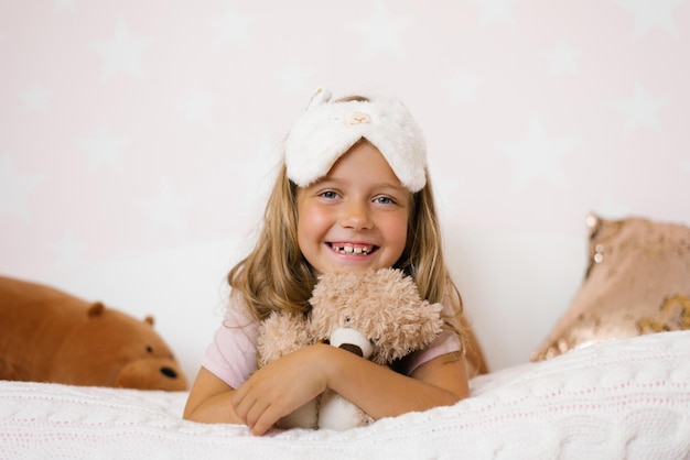 Porträt eines glücklichen kleinen Mädchens mit einer Schlafmaske auf dem Kopf, das auf dem Bett liegt und ihr weiches Spielzeug umarmt