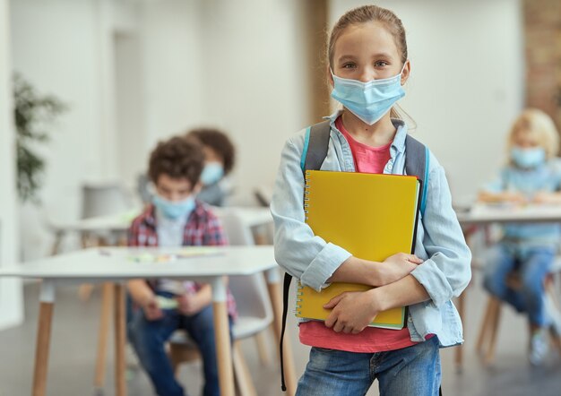 Porträt eines glücklichen kleinen Schulmädchens, das eine Maske trägt, um die Ausbreitung von Covid mit Blick auf die Kamera zu verhindern