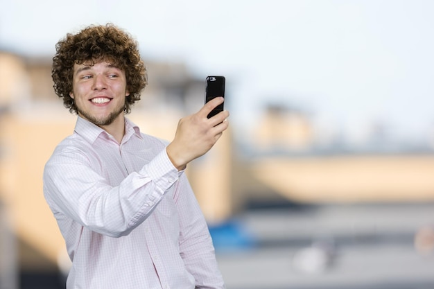 Porträt eines glücklichen lächelnden jungen Mannes mit lockigem Haar, der ein Selfie auf einem verschwommenen Hintergrund macht