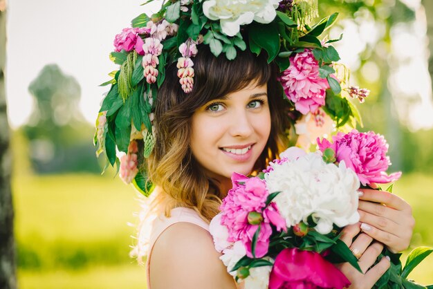 Porträt eines glücklichen lächelnden Mädchens mit einem Kranz von Blumen und einem Blumenstrauß von Pfingstrosen