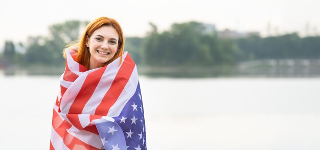 Porträt eines glücklichen lächelnden Mädchens mit USA-Nationalflagge auf ihren Schultern. Positive junge Frau, die den Unabhängigkeitstag der Vereinigten Staaten feiert.