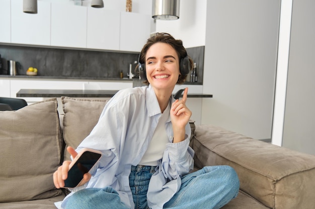 Porträt eines glücklichen Mädchens mit Kopfhörern, das auf dem Sofa tanzt und Musik hört und Spaß in ihrem Haus hat