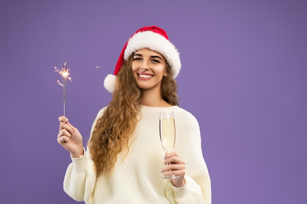Porträt eines glücklichen Mädchens trägt eine Weihnachtsmütze mit einem Glas Champagner und einer Wunderkerze auf violettem Hintergrund