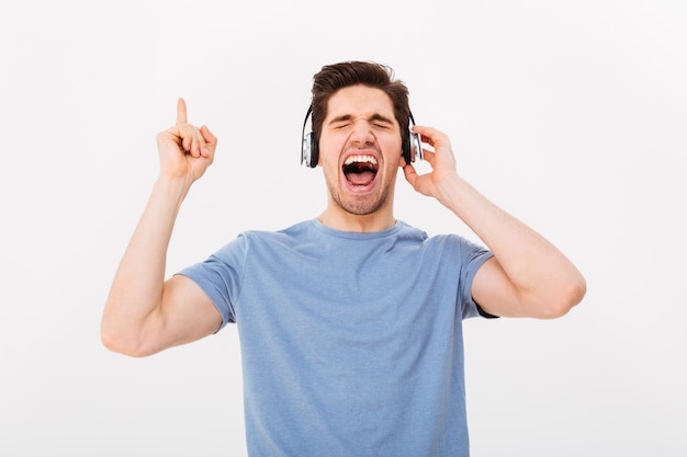 Porträt eines glücklichen Mannes mit kurzen dunklen Haaren, die sein Lieblingslied schreien, während sie Musik über Kopfhörer mit geschlossenen Augen hören, isoliert über weißer Wand