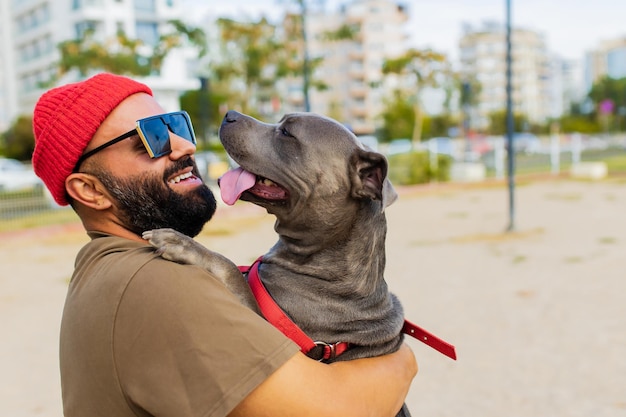 Porträt eines glücklichen Mannes mit rotem Hut und Sonnenbrille mit amerikanischem Terrier im Hundewandergebietspark in der Stadt