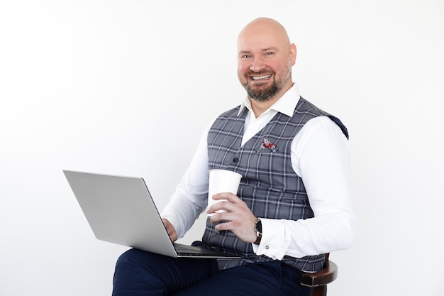 Porträt eines glücklichen Mannes mittleren Alters in grauer Weste, blauer Jeans, weißem Hemd, sitzt mit Laptop und Imbiss