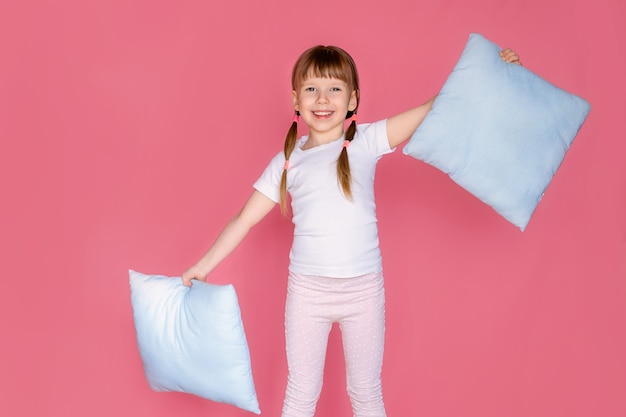 Porträt eines glücklichen niedlichen 5-6 Jahre alten Mädchens, das ihr weiches Kissen umarmt, das Wochenende genießt, sich wohl fühlt, ins Bett geht, weiße Nachtwäsche isoliert über einem rosa Hintergrund anzieht